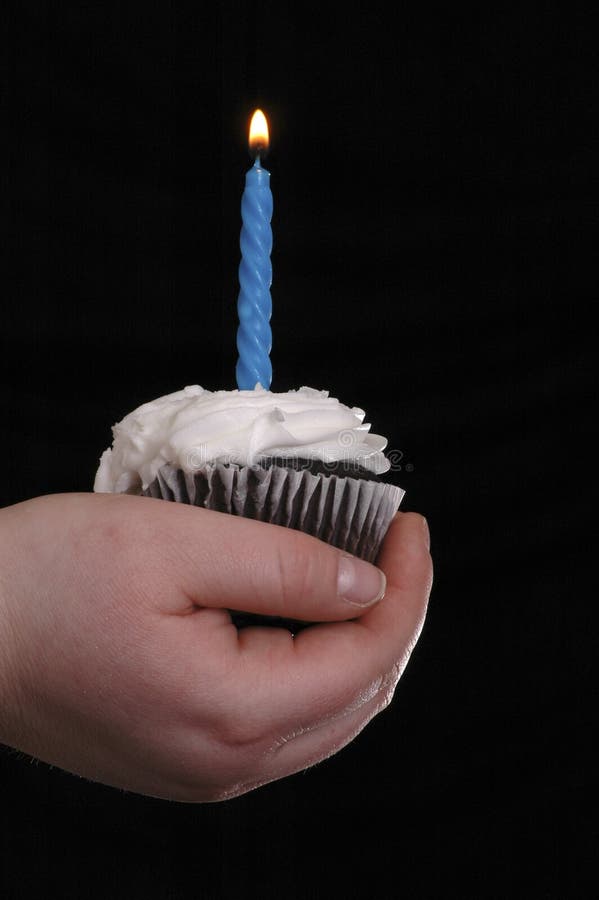 A hand holding a cupcake with one blue candle in it. The candle has been lit. Black background. A hand holding a cupcake with one blue candle in it. The candle has been lit. Black background.