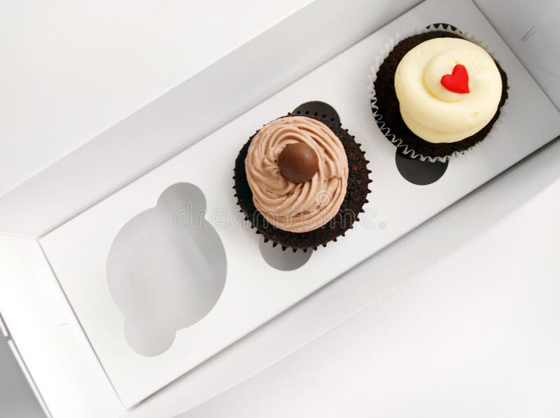 A photograph of two pretty cup cakes in a cardboard box that is made especially for carrying cupcakes and muffins etc for take out or take away. One red velvet cup cake with vanilla cream and a little heart on it to convey love and friendship, one chocolate cupcake with cream. Horizontal color photo, nobody in picture. Happy valentines day gifts or presents. A photograph of two pretty cup cakes in a cardboard box that is made especially for carrying cupcakes and muffins etc for take out or take away. One red velvet cup cake with vanilla cream and a little heart on it to convey love and friendship, one chocolate cupcake with cream. Horizontal color photo, nobody in picture. Happy valentines day gifts or presents.