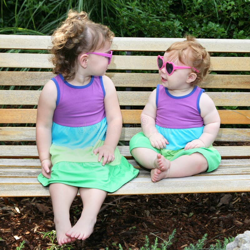 Adorable image of 2 little girls sitting on a park bench in sun dresses and sunglasses. Adorable image of 2 little girls sitting on a park bench in sun dresses and sunglasses