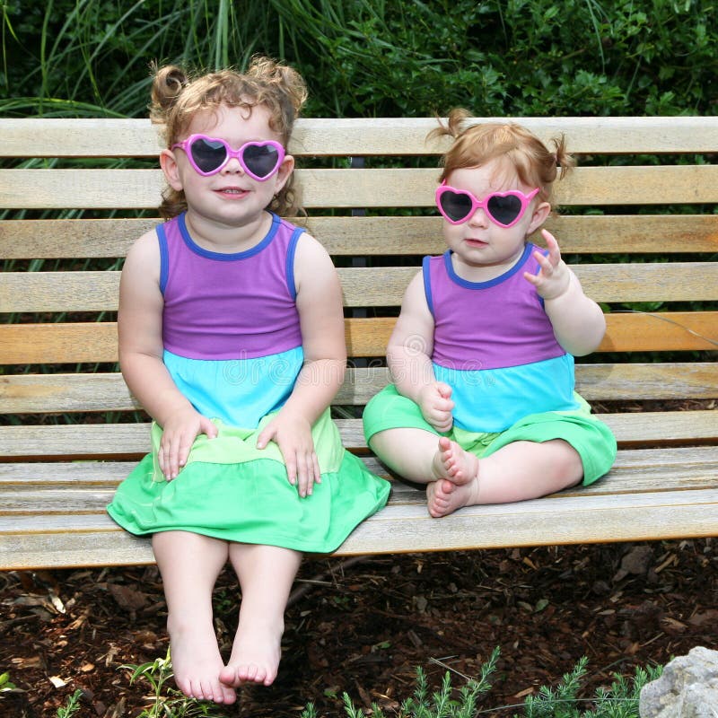 Adorable image of 2 little girls sitting on a park bench in sun dresses and sunglasses. Adorable image of 2 little girls sitting on a park bench in sun dresses and sunglasses