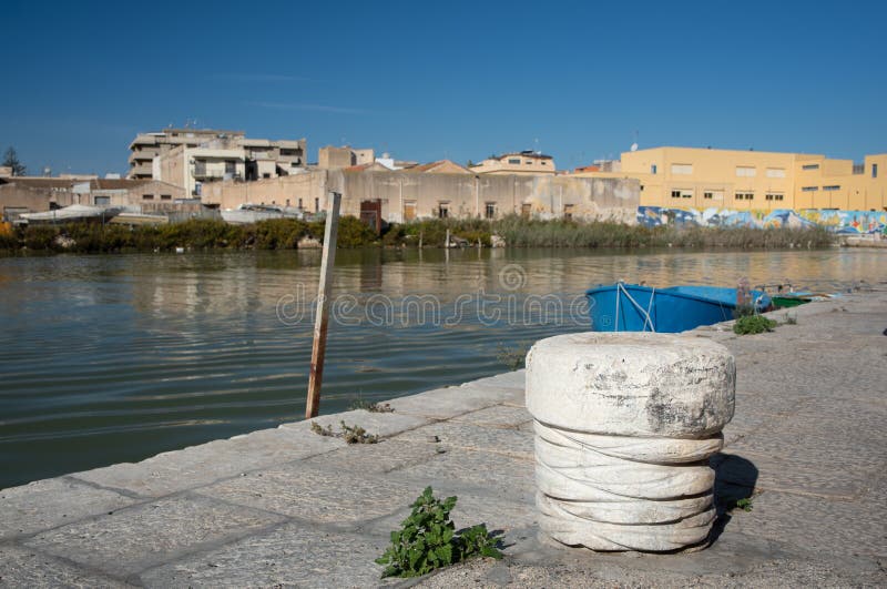 On the bank of a river in Sicily there is a small pillar on the pier to attach ships to. In the background the houses, the river and a small rowing boat, against a blue sky in Italy. On the bank of a river in Sicily there is a small pillar on the pier to attach ships to. In the background the houses, the river and a small rowing boat, against a blue sky in Italy