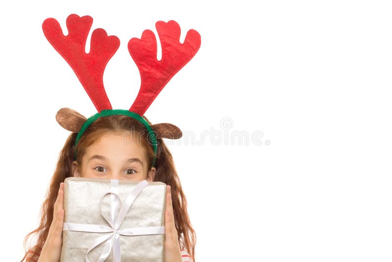 Cropped shot of a little happy girl wearing raindeer Christmas antlers. Cute girl hiding her face behind a gift box looking to the camera from over a present. Cropped shot of a little happy girl wearing raindeer Christmas antlers. Cute girl hiding her face behind a gift box looking to the camera from over a present