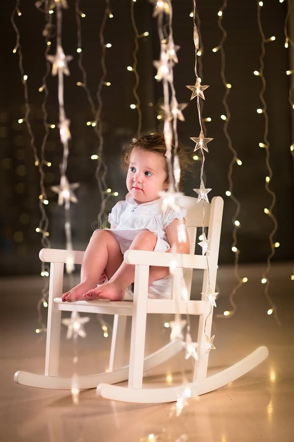 Aanbiddelijk peutermeisje in een witte schommelstoel in donkere ruimte met Kerstmislichten royalty-vrije stock afbeeldingen Aanbiddelijk peutermeisje in een witte schommelstoel in donkere ruimte met Kerstmislichten royalty-vrije stock afbeeldingen