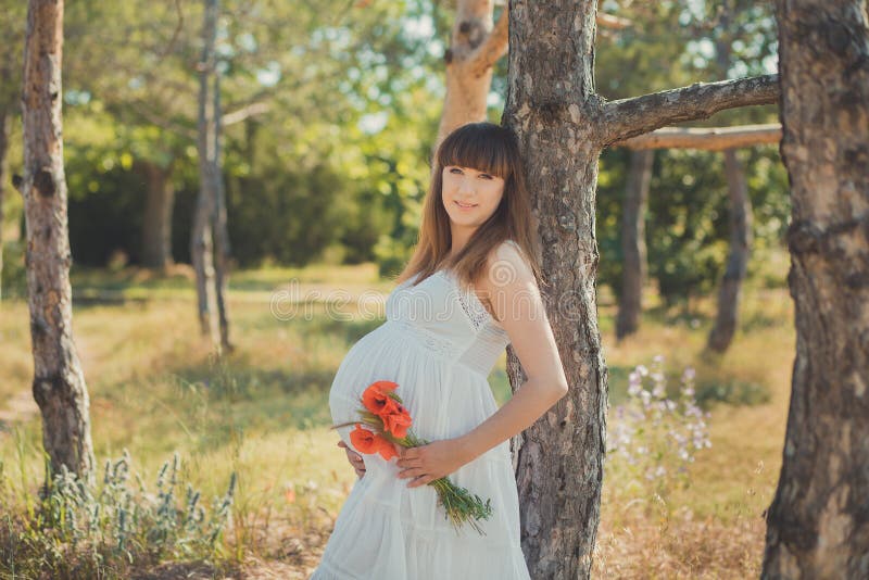 Aanbiddelijke leuke zwangere damevrouw in het witte luchtige kleding stellen dicht bij boom in het bos de buik van de holdingsbui stock foto Aanbiddelijke leuke zwangere damevrouw in het witte luchtige kleding stellen dicht bij boom in het bos de buik van de holdingsbui stock foto