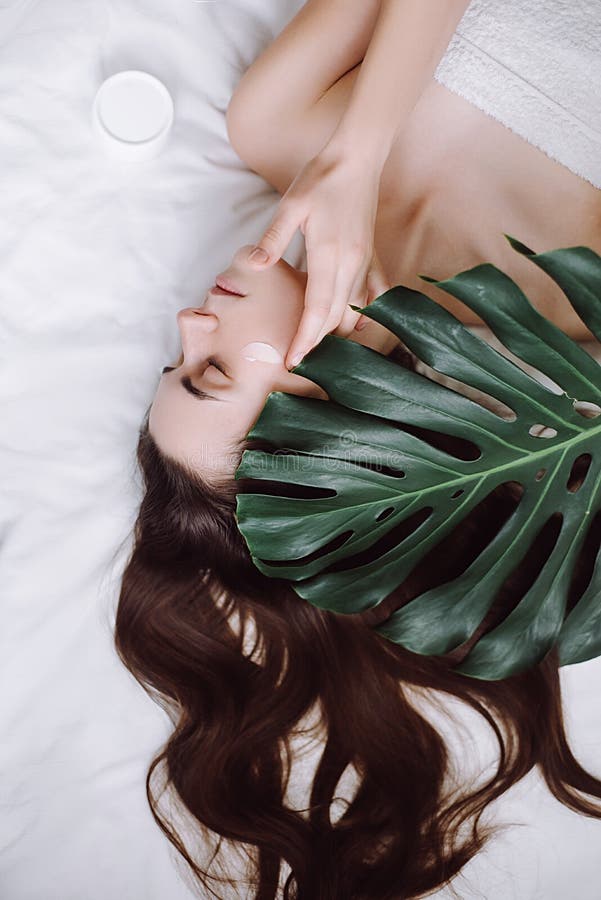 Aantrekkelijk jong langharig meisje die room op haar gezicht toepassen die op het bed met een groen en blad liggen die, day spa g royalty-vrije stock fotografie Aantrekkelijk jong langharig meisje die room op haar gezicht toepassen die op het bed met een groen en blad liggen die, day spa g royalty-vrije stock fotografie