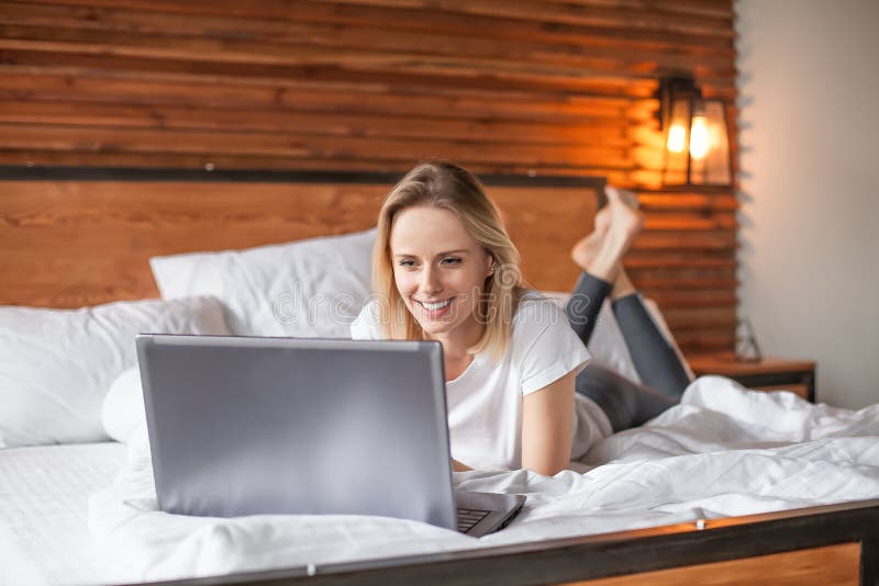 Aantrekkelijke blondevrouw die notitieboekjecomputer met behulp van terwijl het liggen in bed stock afbeelding Aantrekkelijke blondevrouw die notitieboekjecomputer met behulp van terwijl het liggen in bed stock afbeelding