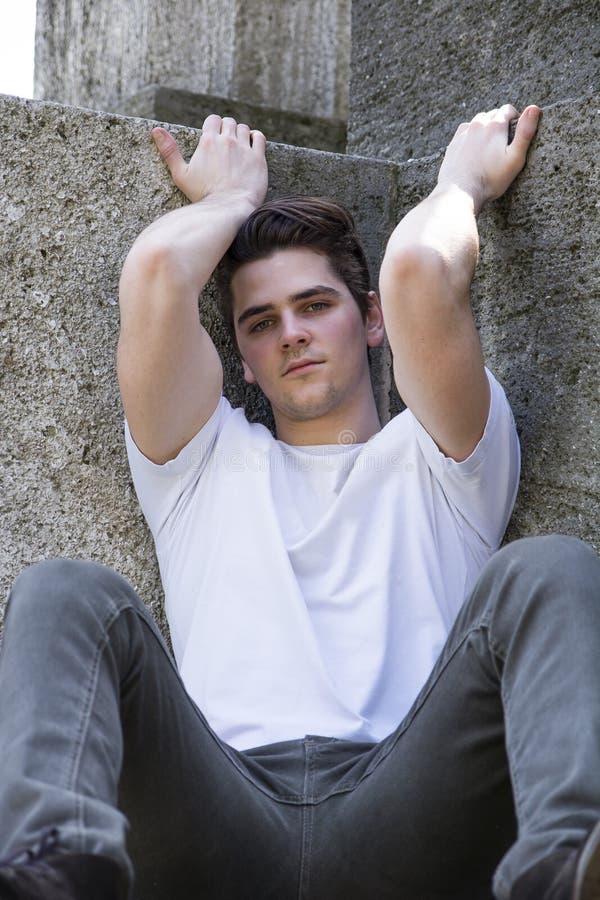 Attractive young man sitting on the ground against stone blocks outdoors, looking at camera, arms up. Attractive young man sitting on the ground against stone blocks outdoors, looking at camera, arms up