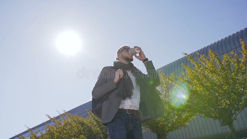 Aantrekkelijke Kaukasische jonge zakenman in jasje met sjaal die die dichtbij de bureaubouw en het drinken koffie lopen