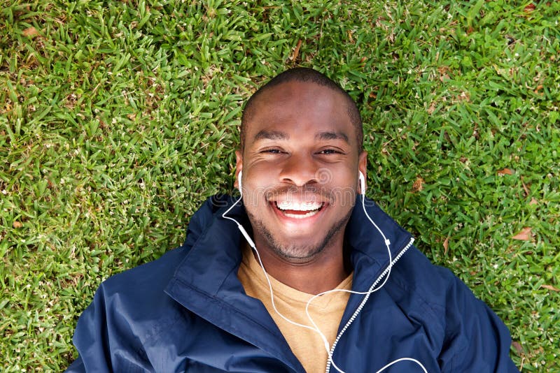 Above of happy black man lying on grass listening to music with earphones