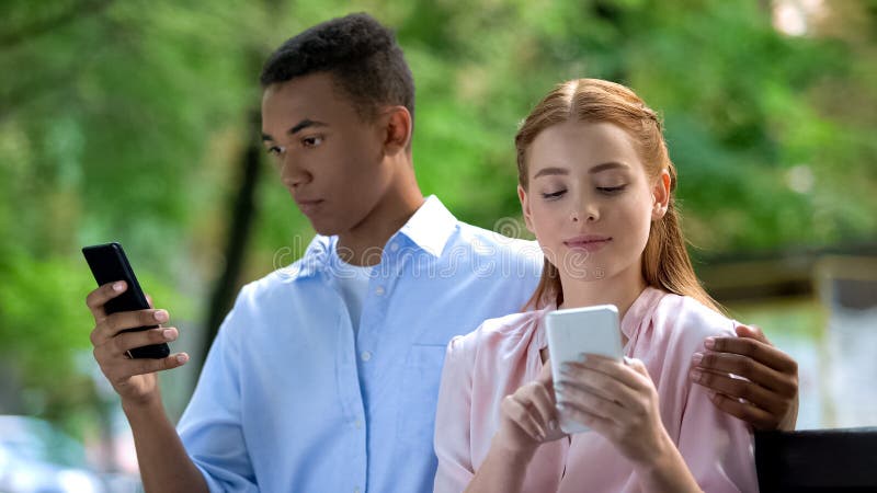 Abrazando a una pareja multiétnica adolescente sentada en un banco en el parque, charlando smartphone foto de archivo libre de regalías