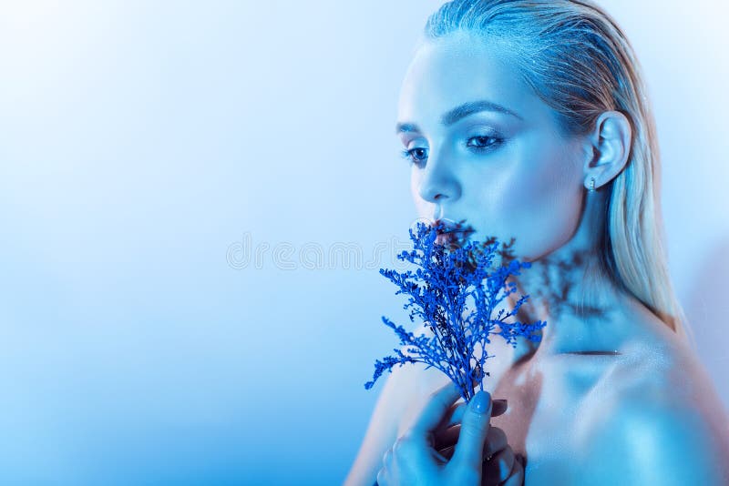 Close up portrait of young beautiful blond model with nude make up, slicked back hair and naked shoulders holding a branch of blue flowers at her face, shadow from them on her neck. Copy-space. Close up portrait of young beautiful blond model with nude make up, slicked back hair and naked shoulders holding a branch of blue flowers at her face, shadow from them on her neck. Copy-space