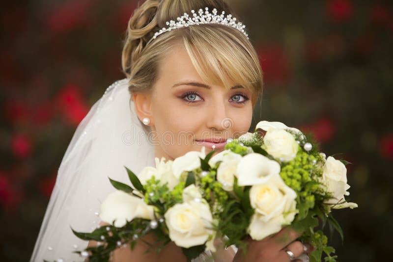 Portrait of pretty attractive young bride with diadem and cute clear blue eyes and perfect skin with nice pearl earring and cute nice lips is holding the bouquet of flowers in front of her face. Portrait of pretty attractive young bride with diadem and cute clear blue eyes and perfect skin with nice pearl earring and cute nice lips is holding the bouquet of flowers in front of her face