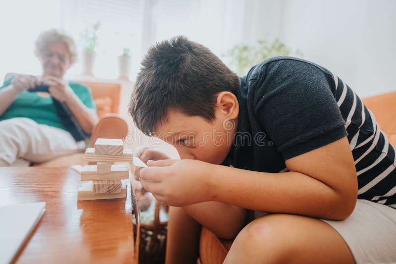 Abuela tejiendo mientras el sobrino juega con bloques de construcción en casa. Un niño se centra intensamente en los bloques de construcción mientras que su abuela tiñe en el fondo capturando un momento familiar acogedor en casa.