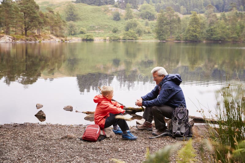 Abuelo y nieto que se sientan en la orilla de un lago que habla, visión elevada, distrito del lago, Reino Unido imagen de archivo libre de regalías Abuelo y nieto que se sientan en la orilla de un lago que habla, visión elevada, distrito del lago, Reino Unido imagen de archivo libre de regalías
