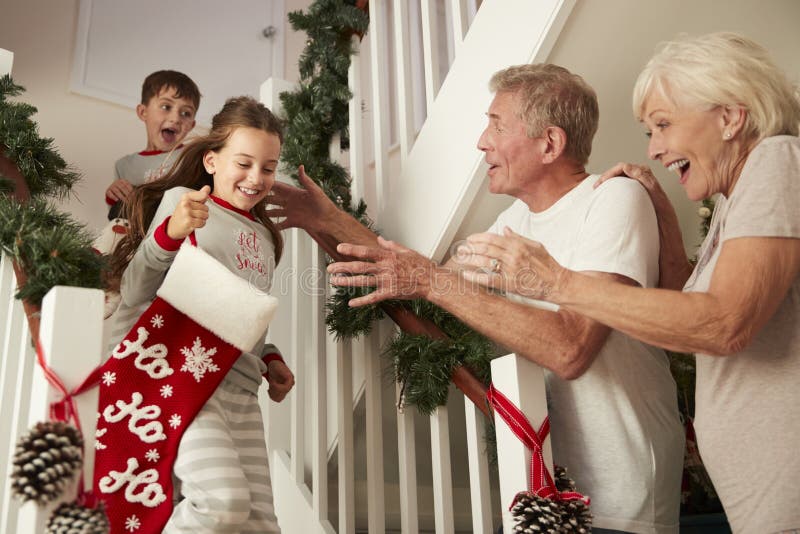Abuelos que saludan a los nietos emocionados que llevan los pijamas que corren abajo de las escaleras que sostienen medias el mañ fotos de archivo Abuelos que saludan a los nietos emocionados que llevan los pijamas que corren abajo de las escaleras que sostienen medias el mañ fotos de archivo