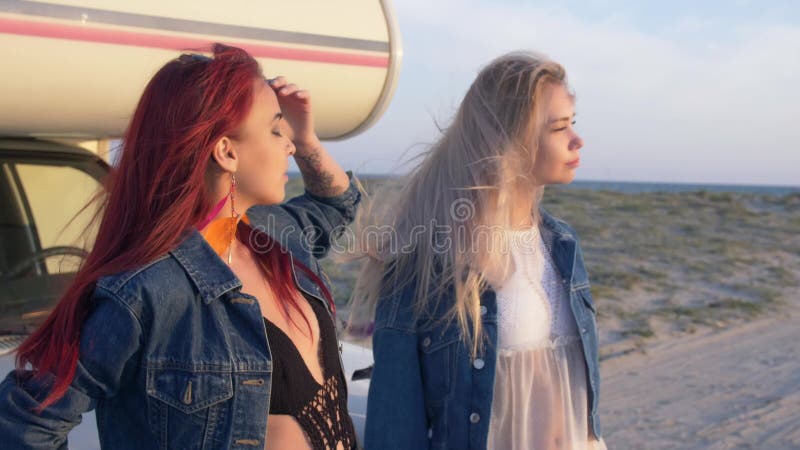 Accoppi delle giovani donne sta sedendosi sul paraurti e godono del tramonto alla spiaggia sabbiosa con la strada