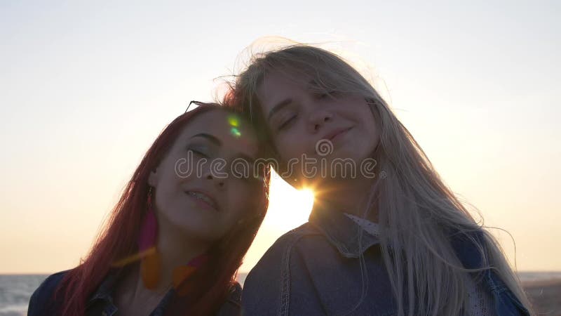 Accoppi delle giovani donne teste a testa in godono del vento del tramonto soffia i capelli alla spiaggia sabbiosa espongono al s