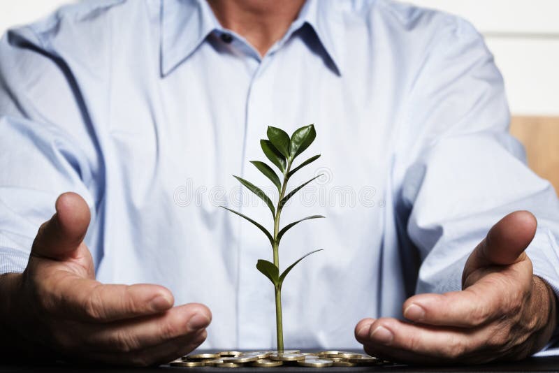 Businessman in blue shirt protecting with hands green plant growing out of coins pile symbolizing safe growth of financial wealth. Businessman in blue shirt protecting with hands green plant growing out of coins pile symbolizing safe growth of financial wealth.