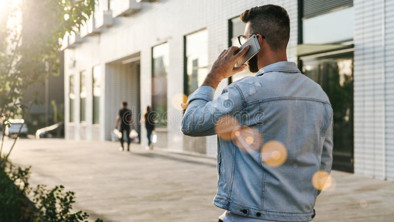 Achter mening De Hipsterzakenman met baard, in denimjasje en in glazen loopt rond stad en vraag op telefoon royalty-vrije stock afbeeldingen Achter mening De Hipsterzakenman met baard, in denimjasje en in glazen loopt rond stad en vraag op telefoon royalty-vrije stock afbeeldingen