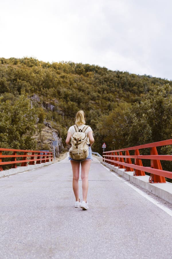 Achteraanzicht jonge blonde vrouw met rugzak op de weg over een brug bij de berg Reis- en avontuurconcept royalty-vrije stock fotografie Achteraanzicht jonge blonde vrouw met rugzak op de weg over een brug bij de berg Reis- en avontuurconcept royalty-vrije stock fotografie