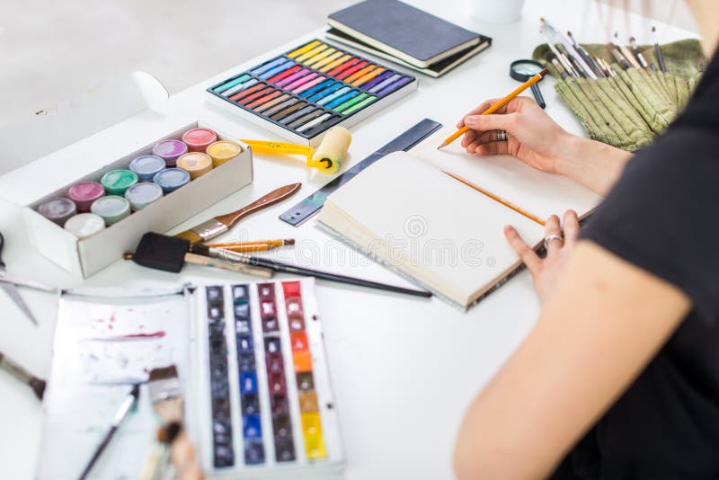 Achterhoekmening van een kunstenaar die nieuw beeld creëren die potloden, pastelkleur, verven en borstels in sketchbook gebruiken
