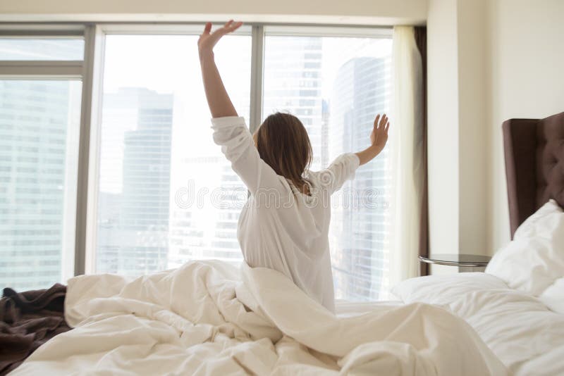 Rear view at woman sitting on bed stretching awaking after sleeping well in the good weekend morning waking up early and happy concept at home or in luxury modern hotel feeling fresh and rested. Rear view at woman sitting on bed stretching awaking after sleeping well in the good weekend morning waking up early and happy concept at home or in luxury modern hotel feeling fresh and rested.