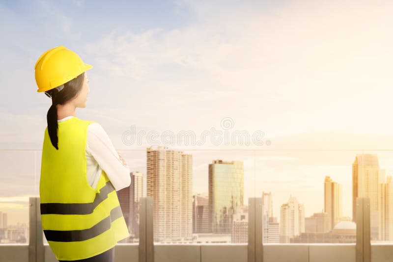 Achtermening van Aziatische arbeidersvrouw in veiligheidsvest, handschoenen, gele helm en beschermend masker die zich bij modern royalty-vrije stock afbeeldingen Achtermening van Aziatische arbeidersvrouw in veiligheidsvest, handschoenen, gele helm en beschermend masker die zich bij modern royalty-vrije stock afbeeldingen