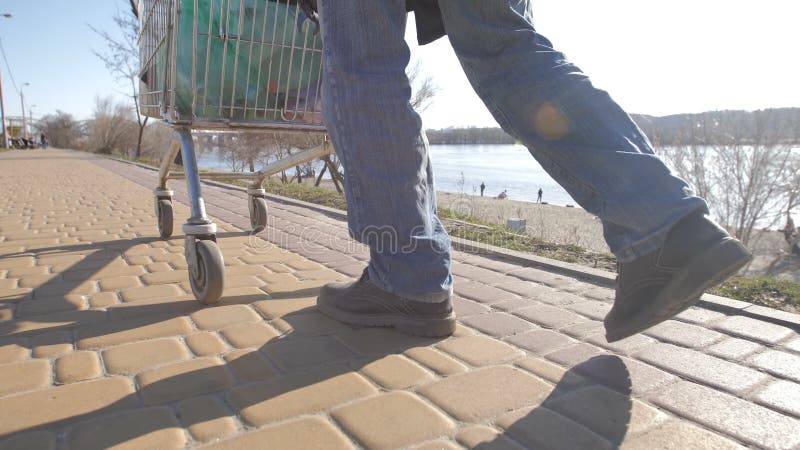 Achtermening van dakloze mensen` s benen die met kar lopen