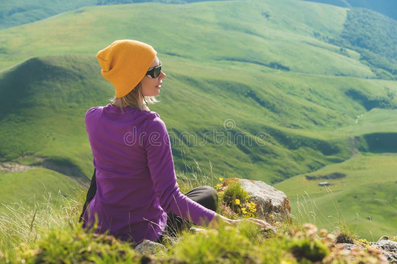 Achtermening van een aantrekkelijke vrouwelijke toeristenzitting op een grasrijke heuvel die zonnebril dragen Binnen genietend va stock foto's Achtermening van een aantrekkelijke vrouwelijke toeristenzitting op een grasrijke heuvel die zonnebril dragen Binnen genietend va stock foto's
