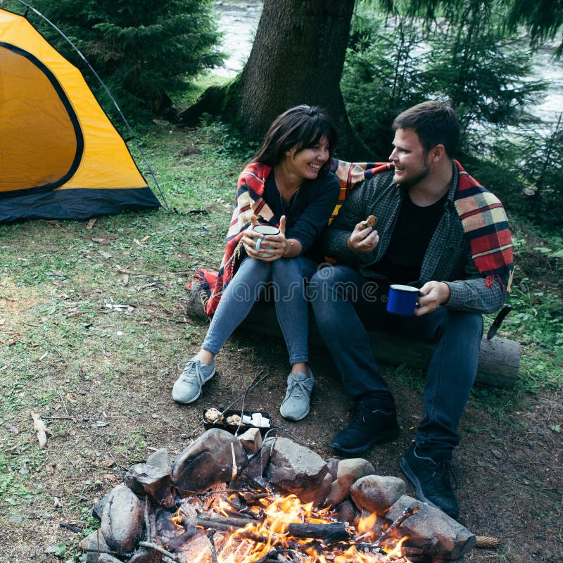 Acople o assento perto do fogo do acampamento e do chá bebendo e dizer histórias barraca e suv no fundo imagens de stock Acople o assento perto do fogo do acampamento e do chá bebendo e dizer histórias barraca e suv no fundo imagens de stock