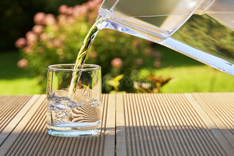 Pouring clear filtered water from a water filtration jug into a glass on the green summer garden background in a warm sunny summer day. Pouring clear filtered water from a water filtration jug into a glass on the green summer garden background in a warm sunny summer day