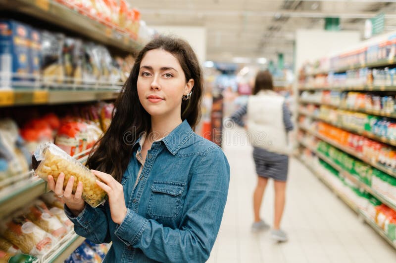 Shopping at the grocery store. Young beautiful woman in denim shirt holding a pack of pasta. In the background, the trading floor of the store. Close up. Shopping at the grocery store. Young beautiful woman in denim shirt holding a pack of pasta. In the background, the trading floor of the store. Close up.