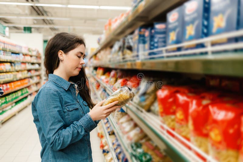 Shopping at the grocery store. A young woman in a denim shirt reads the information on a packet of pasta. In the foreground blurred shelves with products. In profile. Shopping at the grocery store. A young woman in a denim shirt reads the information on a packet of pasta. In the foreground blurred shelves with products. In profile.