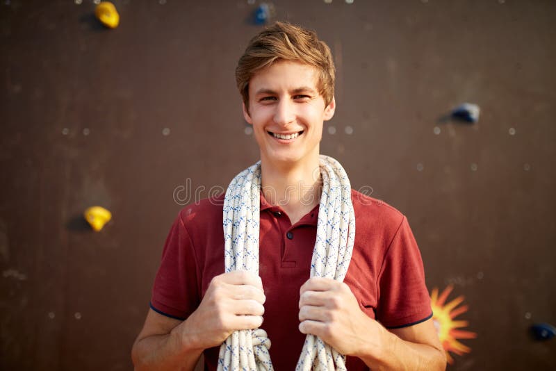 Active young man in sportswear standing with rope on shoulders against artificial training climbing wall. Smiling royalty free stock photo Active young man in sportswear standing with rope on shoulders against artificial training climbing wall. Smiling royalty free stock photo