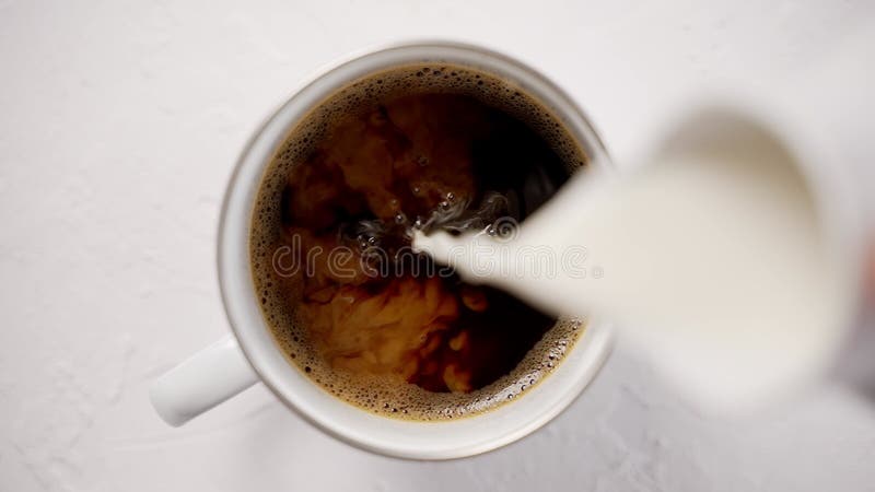 Adding cream to a cup with black, hot coffee placed on white table. Top view, slow motion video. Hot cafe video