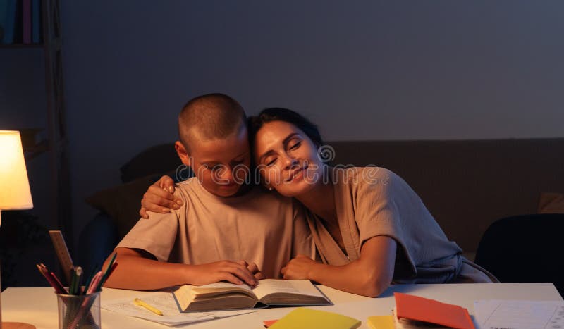 Adolescenta baiat este atent face temele fiul citind o carte mama sta langa el si imbratisare fiul cu zambet. Conceptul educației stock image
