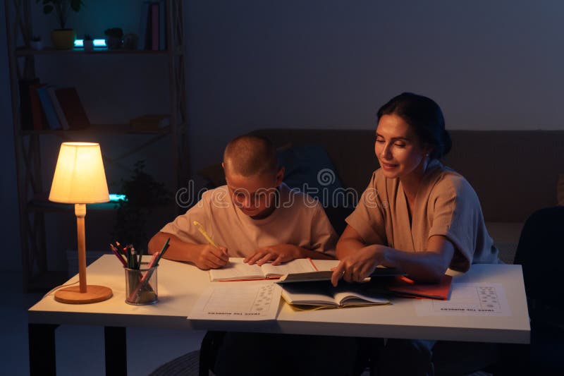 Adolescenta baiat este atent face temele sale în notebook mama deține o carte sits lângă fiu și ajuta cu atribuire. Concept royalty free stock image