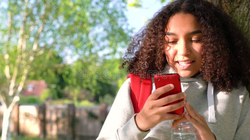 Adolescente afro-americano da menina da raça misturada que inclina-se contra uma árvore usando uma câmera do telefone celular par