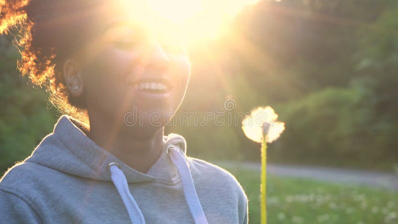 Adolescente afro-americano ou jovem mulher da menina da raça misturada que riem, sorrindo e fundindo um dente-de-leão no por do s