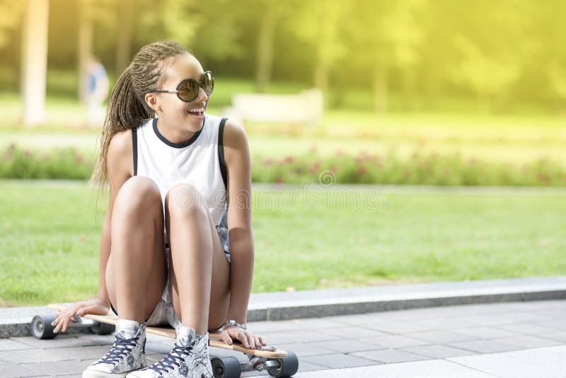 Adolescente afroamericano felice positivo del ritratto con i Dreadlocks che posano su Longboard nell'area del parco fotografia stock libera da diritti