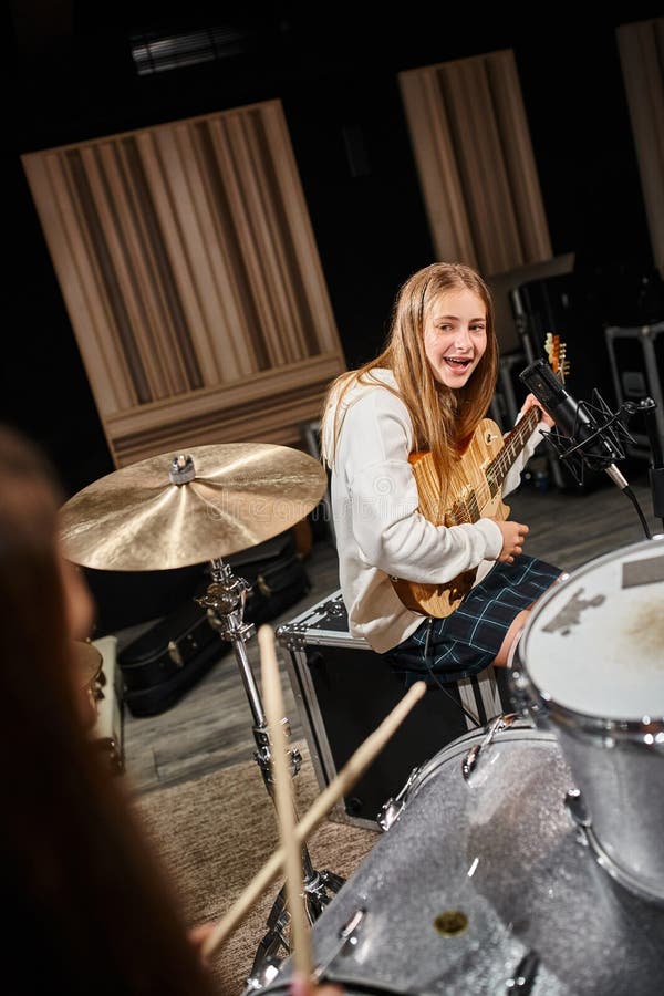 jolly teenage girl in casual outfit playing guitar and looking at her friend playing drums, stock photo. jolly teenage girl in casual outfit playing guitar and looking at her friend playing drums, stock photo