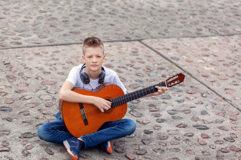 Adolescente con la chitarra acustica e le cuffie che si siedono nel parco immagini stock Adolescente con la chitarra acustica e le cuffie che si siedono nel parco immagini stock