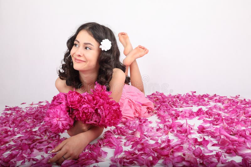 Smiling teenage girl - schoolkid with white flower in brunette hair dressed in pink dress holding bouquet of peonies lying on petals of pink peonies. Pink innocence concept. Smiling teenage girl - schoolkid with white flower in brunette hair dressed in pink dress holding bouquet of peonies lying on petals of pink peonies. Pink innocence concept.