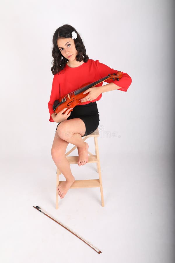 Teenage girl - fashionable schoolkid with brunette hair dressed in red blouse and black skirt sitting on wooden stairs and holding violin as baby. Music and musical instrument concept. Teenage girl - fashionable schoolkid with brunette hair dressed in red blouse and black skirt sitting on wooden stairs and holding violin as baby. Music and musical instrument concept.