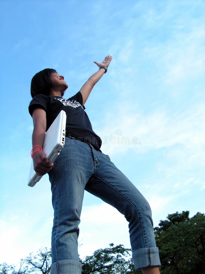 A malay lady wearing jeans and t-shirt, posing with a laptop. Upward shot to show funk and spunk. Fiery blue-ice sky emphasizes her clothes. Can do spirit. Smiling and stretching hand upward to the future. A malay lady wearing jeans and t-shirt, posing with a laptop. Upward shot to show funk and spunk. Fiery blue-ice sky emphasizes her clothes. Can do spirit. Smiling and stretching hand upward to the future.