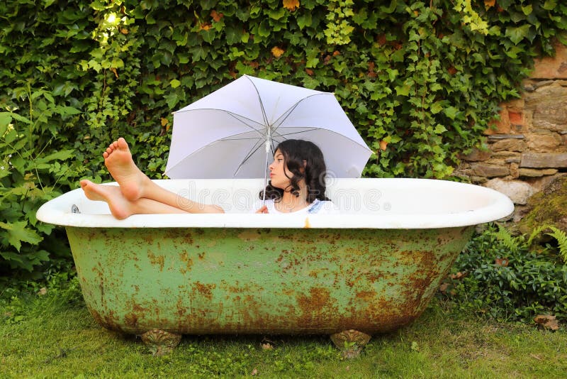 Teenage girl - barefoot schoolkid lying in an old bathtub with bare feet out holding white umbrella. Outdoor with wall overgrown with ivy behind. Retro home related portrait concept. Teenage girl - barefoot schoolkid lying in an old bathtub with bare feet out holding white umbrella. Outdoor with wall overgrown with ivy behind. Retro home related portrait concept.