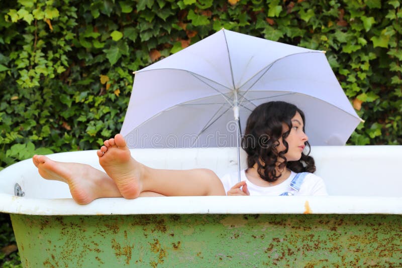 Teenage girl - barefoot schoolkid lying in an old bathtub with bare feet out holding white umbrella. Outdoor with wall overgrown with ivy behind. Retro home related portrait concept. Teenage girl - barefoot schoolkid lying in an old bathtub with bare feet out holding white umbrella. Outdoor with wall overgrown with ivy behind. Retro home related portrait concept.