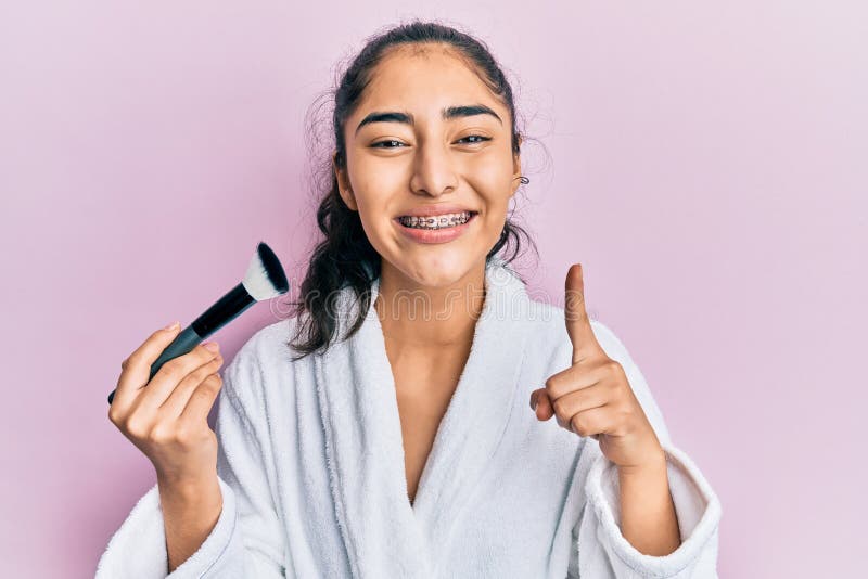 Adolescente hispana con tirantes dentales con bata sosteniendo un cepillo de maquillaje sonriendo con una idea o pregunta señaland. Adolescente hispana con tirantes dentales usando bata sosteniendo un cepillo de maquillaje sonriendo con una idea o pregunta señalando el dedo con la cara feliz número uno