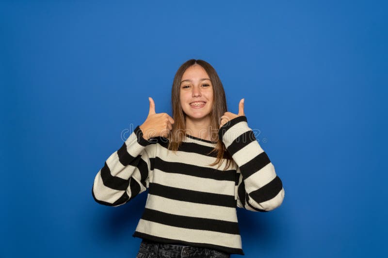 Adolescente ispanico di circa 15 anni con un maglione a strisce che sta sopra un muro blu isolato con i pollici in alto perché immagine stock
