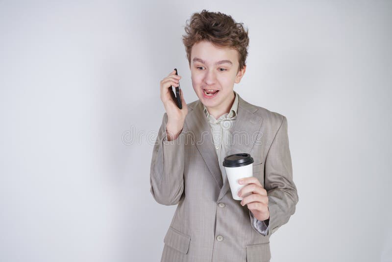 Adolescente joven con emociones sorprendidas en la ropa gris del negocio que se coloca con el tel?fono m?vil y la taza de caf? de foto de archivo libre de regalías Adolescente joven con emociones sorprendidas en la ropa gris del negocio que se coloca con el tel?fono m?vil y la taza de caf? de foto de archivo libre de regalías